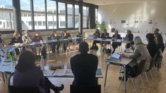 Menschen sitzen in einem sonnigen Saal am Tisch und diskutierren.